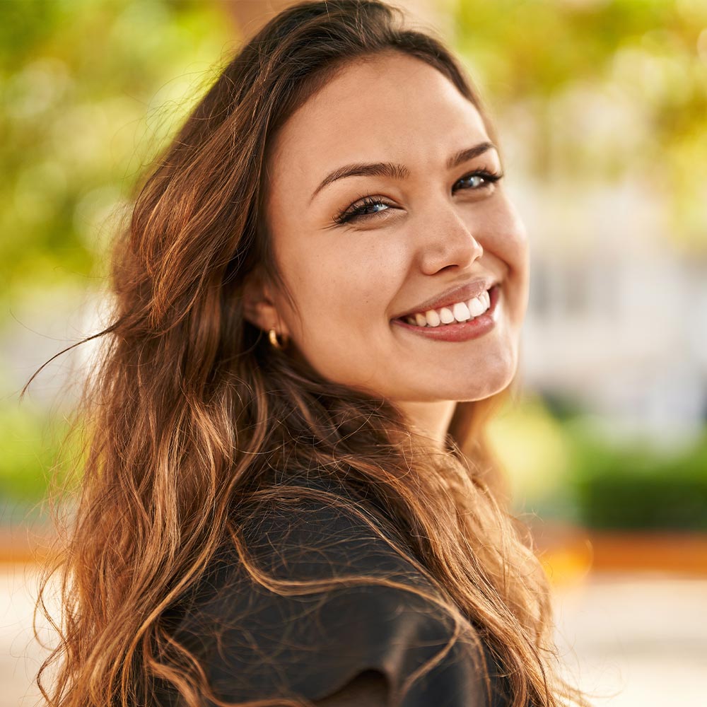 The image features a young woman with long hair smiling at the camera, wearing a dark top, standing outdoors during daylight.