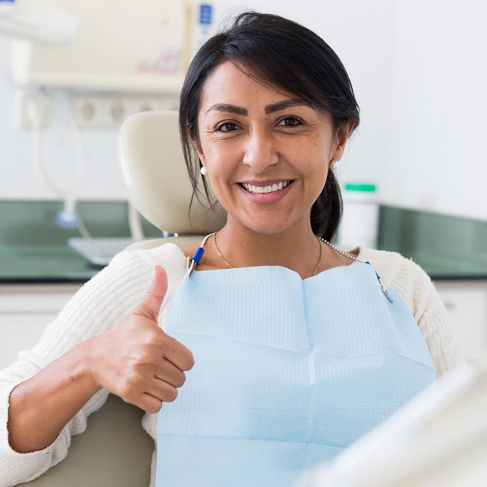 The image shows a woman giving a thumbs up while seated in a dental chair with a mouthguard on, smiling at the camera.