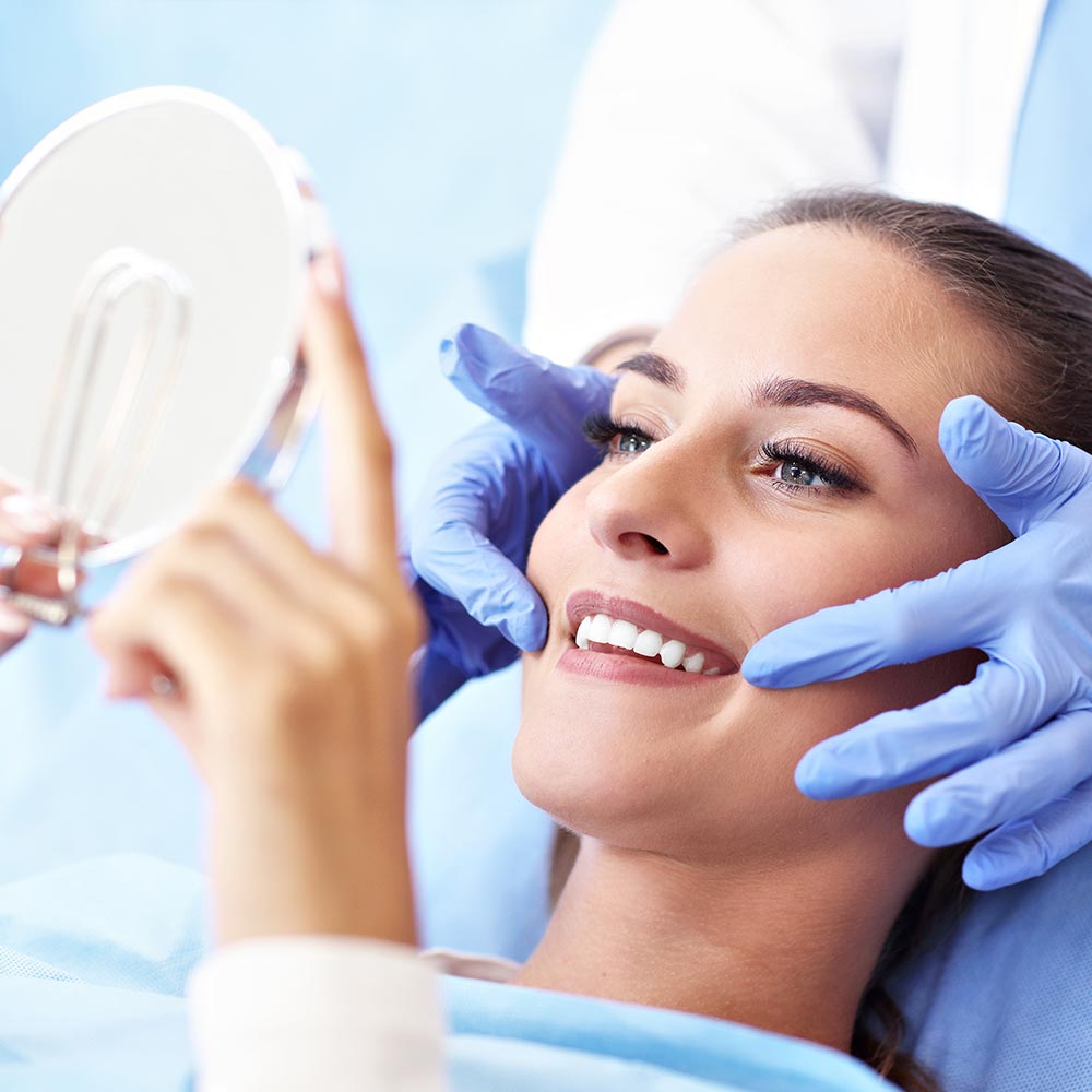 This image shows a person sitting in a dental chair with a smiling expression, looking at the camera, while holding onto a mirror. The individual has their eyes closed and appears relaxed. They are wearing a blue surgical gown, which suggests that they might be undergoing a dental procedure or consultation. A dentist is adjusting a device on the person s face, indicating a professional dental setting.